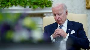 President Joe Biden sitting int he Oval Office with his hands clapsed together and his head bowed down;